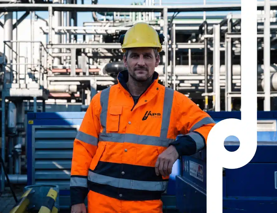 Monteur in oranje hi-vis jas met APS-logo en gele veiligheidshelm voor een industriële procesinstallatie met leidingwerk